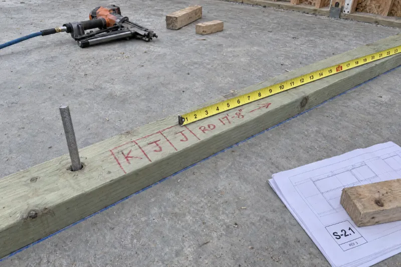 PT 3x6 sill plate on the slab: a hold-down anchor rod beside the king mark, the king and two jacks marked K, J, J, the patio door opening marked RO 17 feet 8 inches in crayon, and a tape hooked on a nail at the last jack line.